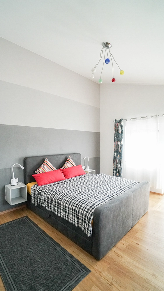 „Hahns Herberge“ – Detailreiche Interioraufnahme; Schlafzimmer in rot und grau gehalten Hochformat Aufnahme mit bunter Lampe auf der Decke.